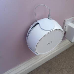 Op de afbeelding: Een witte Deco mesh Wi-Fi-systeem, met een zilveren rand, is gemonteerd op een roze muur. Het apparaat heeft een cirkelvormig ontwerp met een klein, cirkelvormig bovendeel. Een netsnoer is aan de bovenkant aangesloten. Het woord "deco" staat op de voorkant.