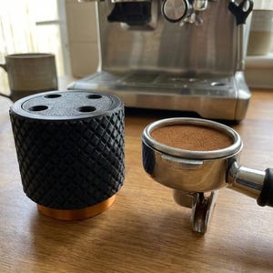 Könnte beinhalten: Ein schwarzer Kaffeetampfer mit strukturiertem Muster und Kupferbasis neben einem Siebträger mit Kaffeepulver. Eine Espressomaschine und eine Keramiktasse im Hintergrund. Die Gegenstände befinden sich auf einer Holzoberfläche.