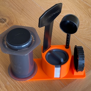 May include: An Aeropress coffee maker set, comprising a clear and grey brewing chamber, a black plunger, and an orange stand with a scoop and filter holder. The stand has the text "AEROPRESS" in black.