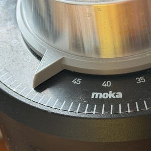 May include: A black coffee grinder adjustment dial featuring white numerical markings from 30 to 45 and the text moka. A grey 3D-printed indicator pointer is attached to the metallic silver top section, aligning with the scale on the black base.