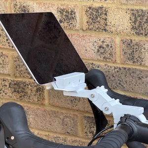 May include: A white tablet holder attached to black bicycle handlebars. The holder is connected to a white adjustable arm. The tablet is black and rectangular. The background is a brick wall.