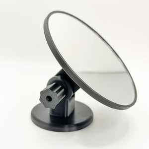 May include: A black, round, adjustable mirror with a black base and a star-shaped knob. The mirror is angled upwards, reflecting the white background. The frame is black.