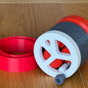 May include: A 3D-printed mechanical device in red, white, grey, and black. It features gears and a crank. A red ring is positioned to the left of the device. The text 'WDT' is printed on the grey band.