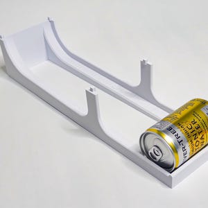 May include: A white plastic can holder with a yellow and silver can of Ver-Tree Tonic Water. The holder has a rectangular base and curved sides with two vertical supports.