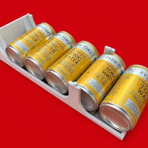 Peut inclure: Un présentoir blanc contenant cinq canettes de Fever-Tree Premium Tonic Water. Les canettes jaunes et argentées sont étiquetées avec le nom de la marque et les détails du produit. Chaque canette contient 150 ml. Le fond est rouge.