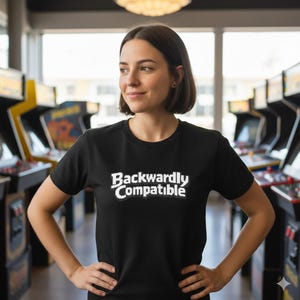 May include: Black crew neck t-shirt with the white text "Backwardly Compatible". The shirt is being worn in front of a row of arcade games.