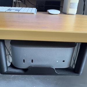 May include: A grey computer is mounted under a wooden desk. The computer is held in place by a black bracket. A white keyboard, mouse, and external hard drive are on the desk.