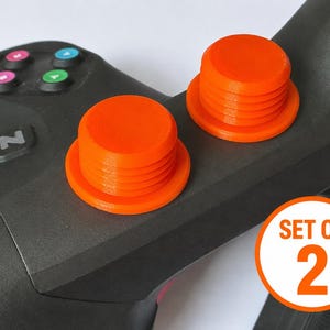 May include: Two bright orange cylindrical button caps with ribbed sides attached to a black gaming controller. The controller features a black button with the letter Z and four smaller buttons in pink, blue, and green. A circular graphic reads SET OF 2.
