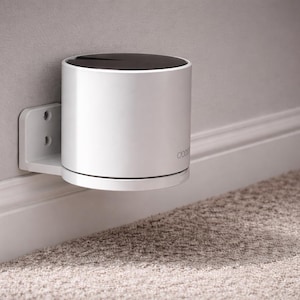 May include: A white, cylindrical device with a black top, mounted on a wall with a white bracket. The device is near a white baseboard and a beige carpeted floor. The word "coova" is printed on the device.