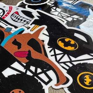 Può includere: Una collezione di adesivi colorati con disegni di monster truck dei cartoni animati. Gli adesivi includono un monster truck marrone con la faccia di un cane, un monster truck nero con il logo di Batman e altri disegni.