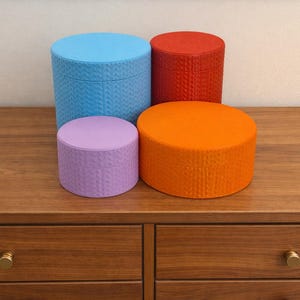 May include: Four colourful, textured storage boxes in blue, red, orange, and lavender sit on a wooden surface. The boxes are cylindrical with matching lids. The wooden surface has a warm, brown tone, and the drawers below have gold knobs.