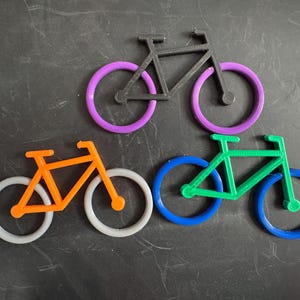 Peut inclure: Trois décorations en forme de vélo aux couleurs vives. L'une est noire avec des roues violettes, une autre est orange avec des roues blanches et la troisième est verte avec des roues bleues. Les vélos sont disposés sur une surface sombre.