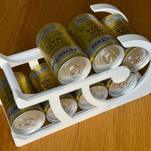 May include: A white, 3D-printed beverage can holder filled with eight cans of Fever-Tree Premium Indian Tonic Water. The cans are gold and silver. The holder is designed to carry the cans in a sleigh-like shape.