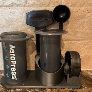 Puede incluir: Juego de cafetera AeroPress negro. El juego incluye una cámara de preparación, una tapa de filtro, una cuchara y un soporte. El logotipo de AeroPress es visible. La cafetera está diseñada para hacer café. Medidas en centímetros.