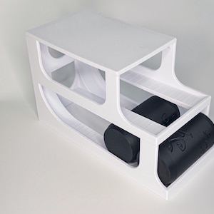 May include: A white, two-tiered wine rack with space for bottles. Two black wine bottles are in the rack. The rack has a modern design with curved cutouts. The rack is made of white material and has a minimalist aesthetic.