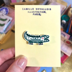 Peut inclure: Une épingle en forme d'alligator verte et blanche avec des accents rouges. L'épingle est sur une carte jaune avec le texte "Camille Devallois Illustration, Paris".