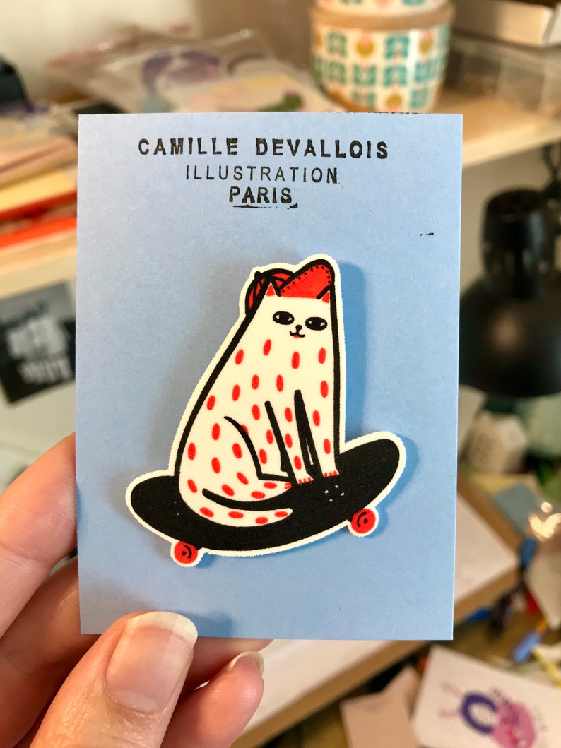 Puede incluir: Un gato blanco con lunares rojos que lleva un sombrero rojo est&aacute; montando una patineta negra con ruedas rojas. El gato est&aacute; mirando hacia la derecha. La imagen est&aacute; sobre un fondo azul con el texto "Camille Devallois Illustration Paris" en la parte superior.
