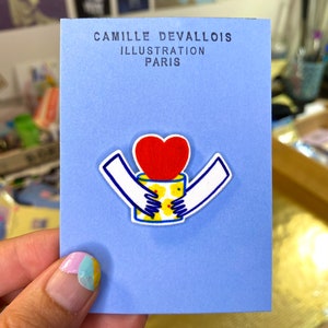 May include: A blue card with white text that reads "Camille Devallois Illustration Paris". A white outline of two hands holding a red heart with a yellow flower pattern inside.