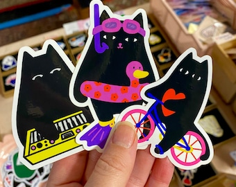 以下が含まれることがあります： 異なるデザインの黒い猫のステッカーが3つあります。1匹の猫はピンクと白のライフジャケットと紫色のゴーグル付きシュノーケルを着用しています。もう1匹の猫はピンクの車輪と青いフレームの自転車に乗っています。3匹目の猫は黄色のキーボードを弾いています。