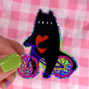 Op de afbeelding: Een zwarte kat met drie ogen die op een glimmende paarse fiets met roze wielen rijdt en een rood hart vasthoudt. De kat en de fiets bevinden zich op een roze-witte ruitjesachtergrond.