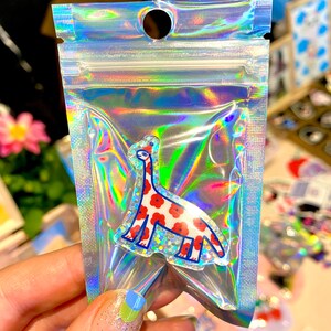 Peut inclure: Un sac en plastique transparent et irisé contenant un autocollant en forme de dinosaure. L'autocollant présente un corps blanc avec des motifs de fleurs rouges et des contours bleus. Le sac est scellé.