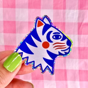 Peut inclure: Un autocollant représentant une tête de tigre bleue et blanche avec un nez rouge et des moustaches. L'autocollant a une finition holographique.