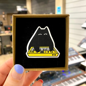Peut inclure: Autocollant d'un chat noir jouant d'un synthétiseur jaune, sur fond noir. L'autocollant est présenté dans une petite boîte carrée en carton. Le chat a des yeux blancs et joue du synthétiseur avec ses pattes.