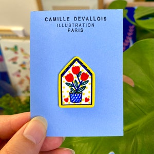 Peut inclure: Une épingle en forme de maison jaune avec une plante en pot bleu et blanc avec des fleurs rouges et des pois jaunes sur un fond blanc. L'épingle indique "Camille Devallois Illustration Paris" au dos.