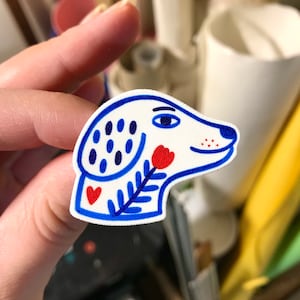 Può includere: Una testa di cane in ceramica bianca con dettagli blu e rossi. Il cane ha un orecchio con un motivo blu e bianco e un fiore rosso sul naso. Gli occhi del cane sono blu e la bocca è leggermente aperta.