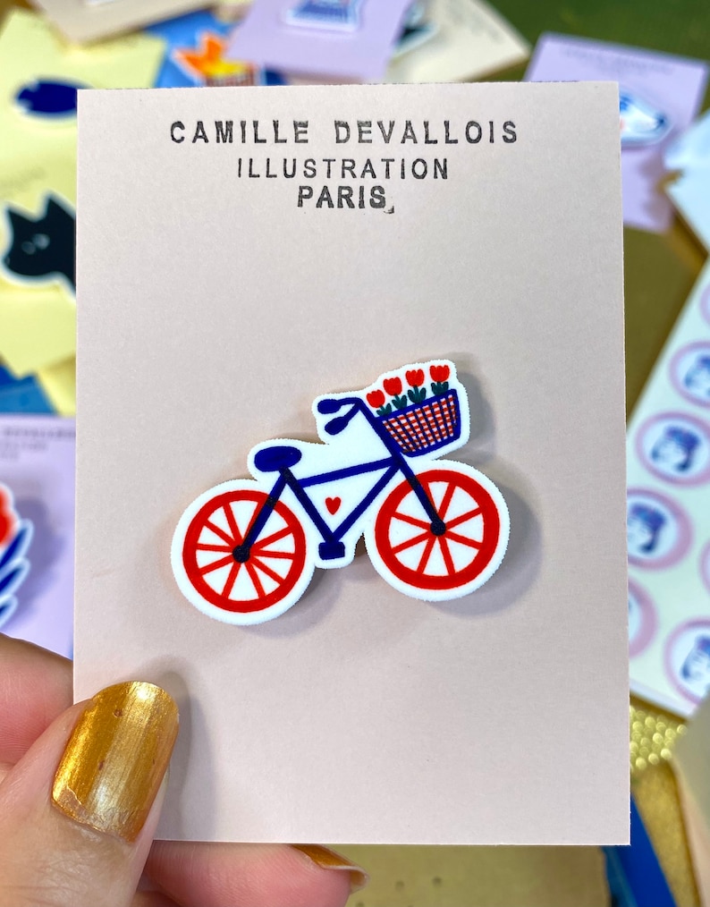 May include: A blue bicycle with red wheels and a basket full of red flowers. The bicycle has a small heart on the frame. The bicycle is on a white card with the text "Camille Devallois Illustration Paris" printed on it.