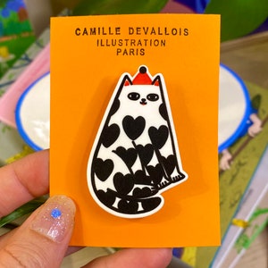 Könnte beinhalten: Eine weiße Katze mit schwarzen Herzen, die auf ihren Körper gedruckt sind, trägt einen roten Hut. Die Katze sitzt auf einer leuchtend orangefarbenen Karte mit dem Text "Camille Devallois Illustration Paris".
