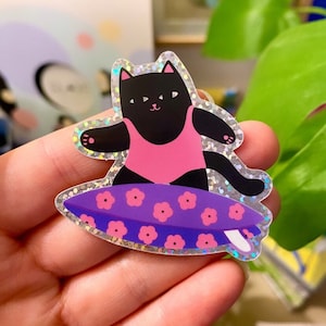 Peut inclure: Un chat noir portant un maillot de bain rose surfe sur une planche de surf violette avec des fleurs roses. L'autocollant est iridescent et a une finition scintillante.