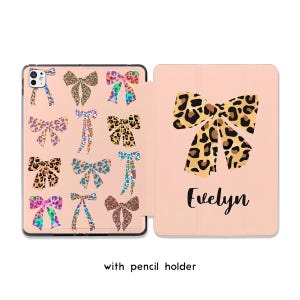 May include: A pink tablet case with a leopard print bow design. The case features multiple bow patterns on the front cover and a large bow with the name "Evelyn" on the back. Includes a pencil holder.