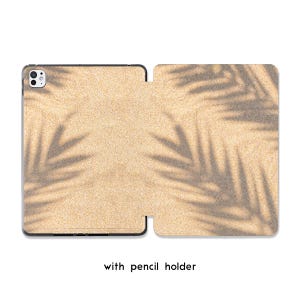 Könnte beinhalten: Eine beige Tablet-Hülle mit schwarzem Rand, die eine sandartige Textur und ein Palmenblatt-Schatten-Design aufweist. Der Text "with pencil holder" ist unten aufgedruckt. Die Hülle ist geöffnet.