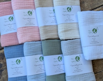 May include: A collection of folded, organic cotton swaddles in various pastel colors, including pink, blue, green, and beige. Each swaddle is wrapped with a white label featuring text and a globe graphic. The swaddles are arranged on a rustic wooden surface.