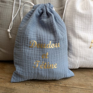 May include: Two drawstring bags made of soft, textured fabric. One bag is light blue with gold lettering that says "Doudou et Tetine". The other bag is white.