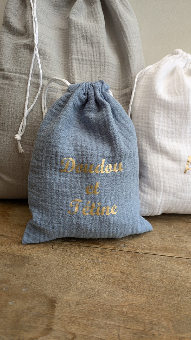 May include: Two drawstring bags made of soft, textured fabric. One bag is light blue with gold lettering that says "Doudou et Tetine". The other bag is white.