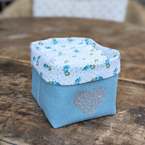 Lot de 12 lingettes format 8 x 8 cm en microfibre et coton imprimé fleurs vertes sur fond écru. Elles sont rangées dans un panier en lin vert avec un coeur pailleté .  Il mesure 9 cm de côté environ.