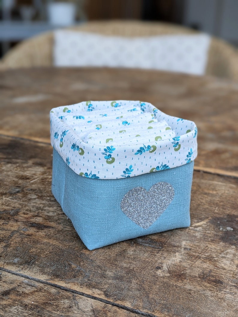 Lot de 12 lingettes format 8 x 8 cm en microfibre et coton imprimé fleurs vertes sur fond écru. Elles sont rangées dans un panier en lin vert avec un coeur pailleté .  Il mesure 9 cm de côté environ.