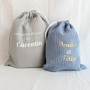May include: Two drawstring bags, one gray and one blue. The gray bag has the text "Les petites affaires de Corentin" in white. The blue bag has the text "Doudou et t&eacute;tine" in gold.
