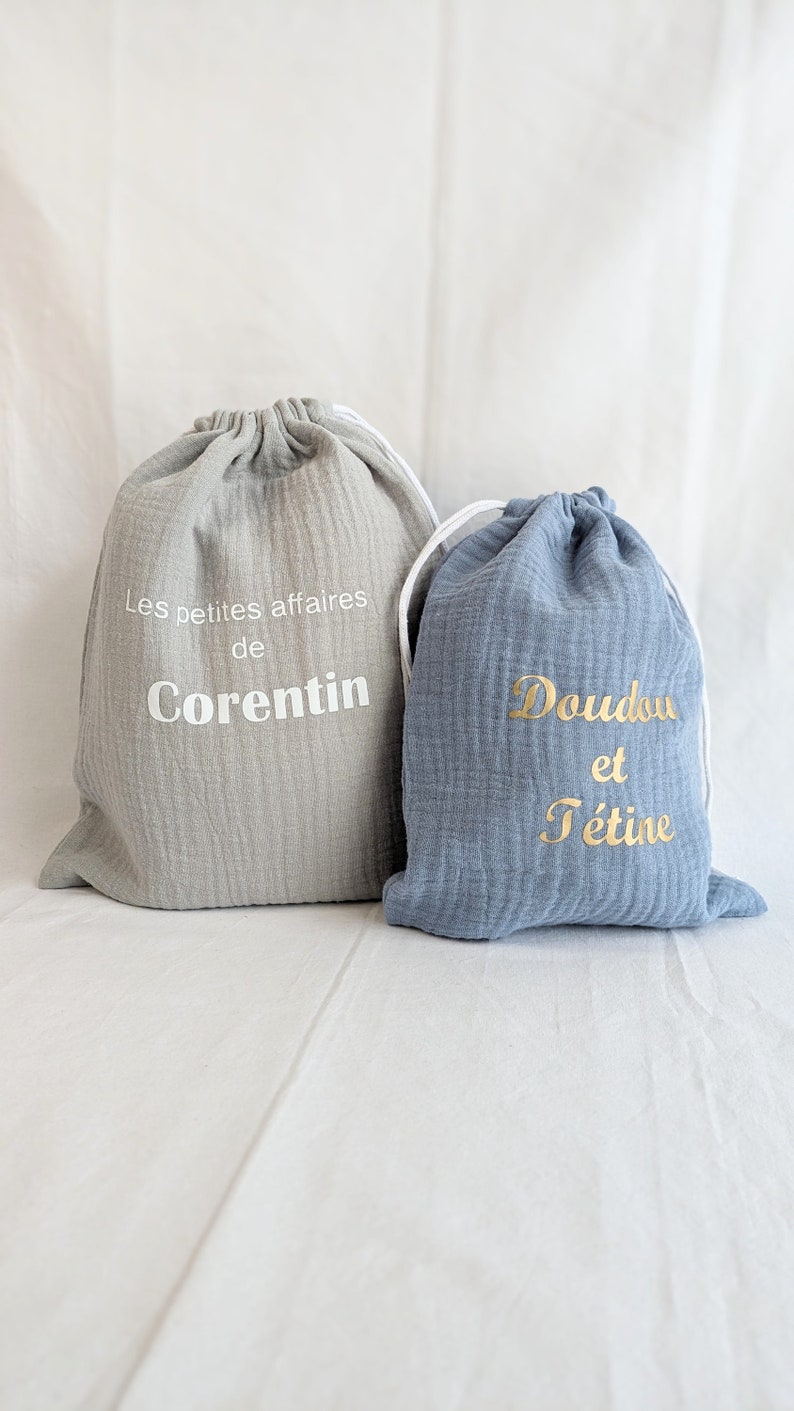 May include: Two drawstring bags, one gray and one blue. The gray bag has the text "Les petites affaires de Corentin" in white. The blue bag has the text "Doudou et t&eacute;tine" in gold.
