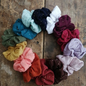 May include: A set of 15 colorful scrunchies with gold glitter accents. The scrunchies are made of a soft, textured fabric and are arranged in a circle on a wooden surface.