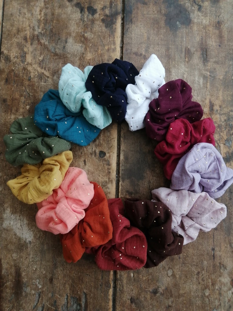 May include: A set of 15 colorful scrunchies with gold glitter accents. The scrunchies are made of a soft, textured fabric and are arranged in a circle on a wooden surface.