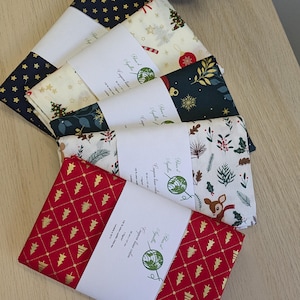 Peut inclure: Cinq coupons de tissu pliés avec des motifs de Noël : étoiles dorées sur fond marine, sapins et boules sur crème, flocons dorés sur vert foncé, feuillage et faon sur blanc, et sapins dorés sur un quadrillage rouge à losanges.