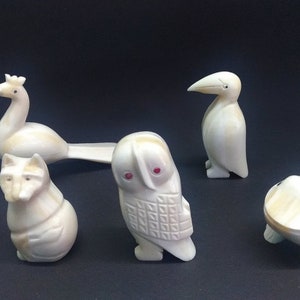 May include: A collection of carved ivory-colored animal figurines. The set includes a peacock, a raccoon, an owl with red eyes, a penguin, and a turtle. Each figurine is detailed and stands on a dark background.