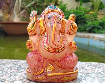May include: A pink and gold statue of the Hindu deity Ganesha, seated with hands raised. The statue has intricate details and is set against a blurred background of greenery and a water feature.
