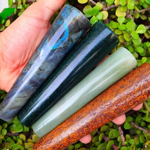 Op de afbeelding: Vier gepolijste stenen pijpen in tinten blauw, groen en oranje worden getoond. De pijpen worden in een hand gehouden en rusten op een bed van groene bladeren. De pijpen hebben unieke patronen en texturen.