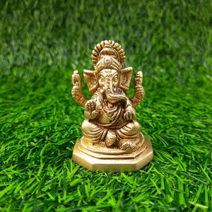May include: A gold-coloured statue of Ganesha, the Hindu deity, in a seated pose. The statue features detailed craftsmanship and is placed on a bed of green artificial grass. Ganesha is a symbol of wisdom and good fortune.