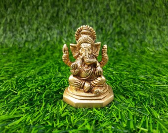 May include: A gold-colored statue of the Hindu deity Ganesha, seated in a meditative pose. The statue has intricate details and is set against a backdrop of green artificial grass. The figure is a symbol of wisdom and prosperity.