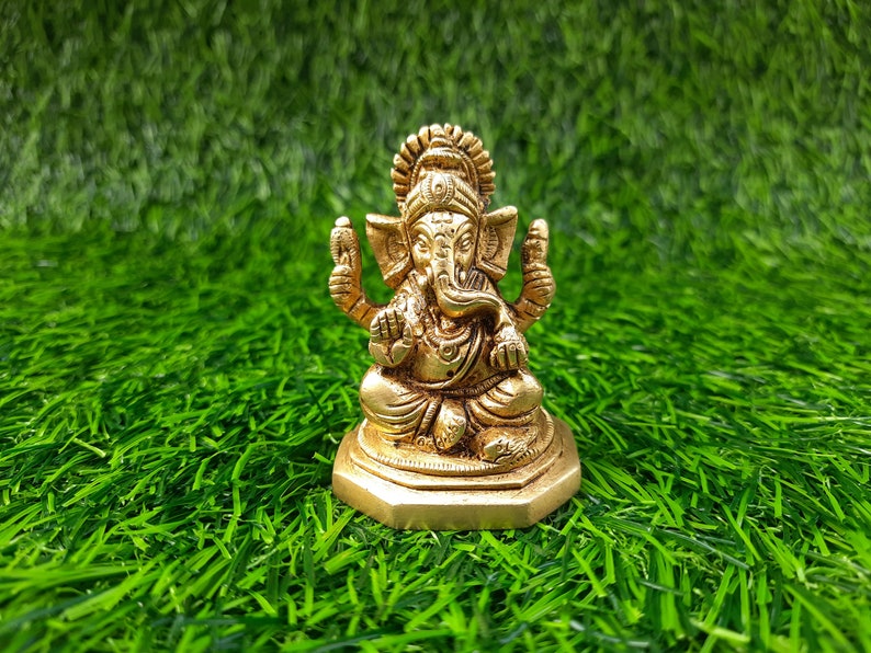May include: A gold-coloured statue of Ganesha, the Hindu deity, in a seated pose. The statue features detailed craftsmanship and is placed on a bed of green artificial grass. Ganesha is a symbol of wisdom and good fortune.